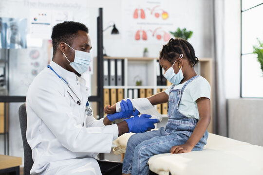 Lovely cute dark-skinned small girl in facial mask sitting on couch in medical office, while handsome professional African male doctor in protective mask applying bandage on her damaged arm