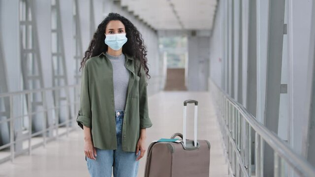 Hispanic girl with curly hair passenger woman wears protective medical mask on female face stands near suitcase in airport terminal railway station looking at camera traveling in pandemic quarantine