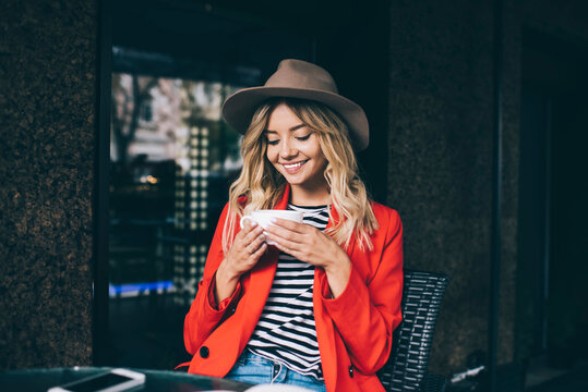 Cheerful hipster girl in stylish hat enjoying leisure pastime in street cafe holding white cup with hot tasty tea and smiling, happy Caucasian female customer 20s spending weekend in city