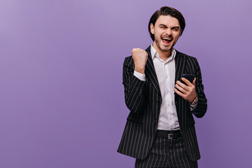 Joyful young gentleman in classic striped suit and light shirt, rejoicing, holding mobile phone and...