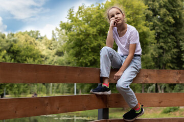 A young girl looks up while sitting on a wooden fence against the backdrop of green trees.