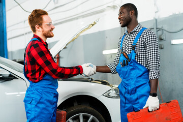 African american man holding toolbox and red-haired mechanics shaking hands while standing against the backdrop of a car in a car service