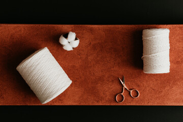 Two rolls of cotton rope, a gold rose scissors and a white cotton flower head on rust and orange color canvas on black background.  