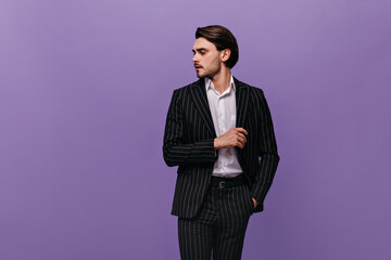 Obraz na płótnie Canvas Handsome young gentleman with brunette hair, white shirt and dark striped suit, putting hand in pocket and looking aside. Man posing isolated over violet background