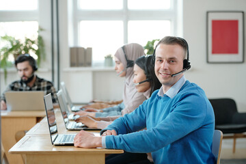 Fototapeta na wymiar Young successful operator with headset looking at you with smile against his co-workers