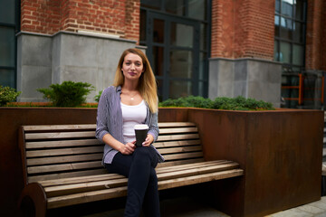 Obraz premium Portrait of attractive female student with disposable takeaway cup resting at city bench and looking at camera during coffee break in campus, beautiful hipster girl with caffeine beverage posing
