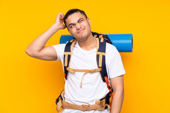 Young mountaineer asian man with a big backpack isolated on yellow background having doubts and with confuse face expression