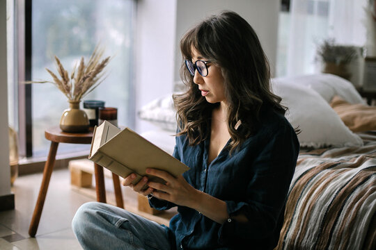 Young focused asian woman wearing eyeglasses reading book while sitting in lotus pose on floor in cozy living room