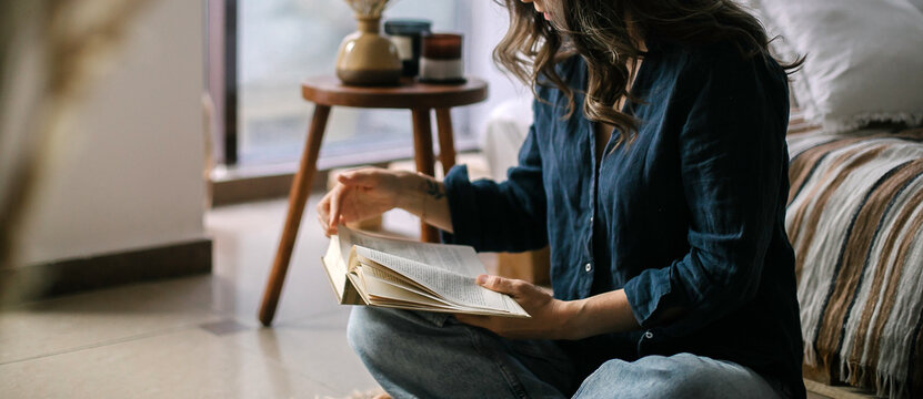 Young woman reading book while sitting in lotus pose on floor in cozy living room, spending leisure time at home