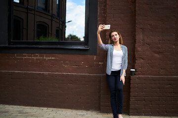 Attractive female blogger in casual clothing looking at front mobile camera while shooting influence video vlog, millennial hipster girl using modern smartphone technology for clicking selfie images