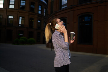 Fototapeta premium Cheerful hipster girl with takeaway coffee to go talking with best friend and laughing during phone conversation in roaming,happy Caucasian woman with caffeine beverage enjoying positive communication