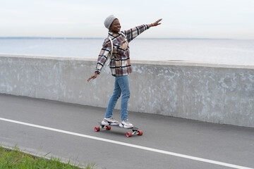Freedom and urban lifestyle concept: young casual girl skateboarding on longboard at city road near sea coast. Happy millennial active african american woman enjoy extreme leisure activity outdoors.