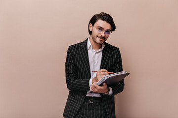 Handsome young gentleman with brunette hair, white shirt, black striped suit smiling, writing something in notebook and looking into camera. Man posing isolated over beige background