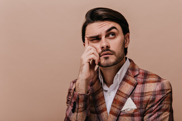 Elegant young brunette with bristle, white collar and plaid blazer making gesture of thinking and raising one eyebrow. Man looking up and posing against beige background