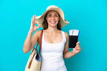 Young caucasian woman holding a beach bag and passport with pamel isolated on blue background showing ok sign with fingers