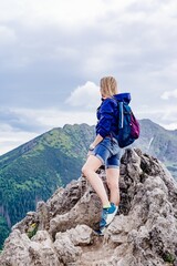 Rear view of a blonde female traveler standing on a rock. She admires the beautiful mountain views.