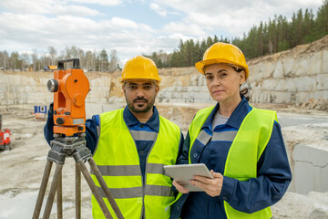 Intercultural colleagues in workwear and hardhats standing by total station