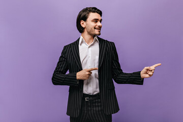 Obraz na płótnie Canvas Joyful young dark-haired gentleman, wearing white shirt and striped suit, looking and pointing aside on copy space isolated over violet pastel background