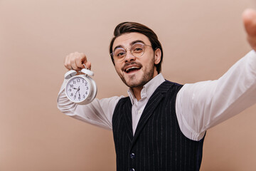 Cheerful young brunette-haired man with glasses, white shirt and black waistcoat, holding clock,...