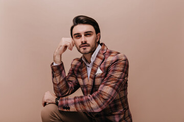 Handsome young man with brunette hair and bristle sitting against beige pastel background and...