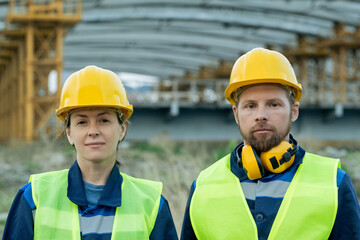 Portrait of two engineers in work helmets looking at camera while working together on construction site