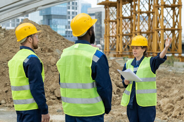 Foreman with blueprint pointing at construction and talking to workers during their work outdoors