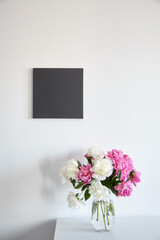 Black canvas mockup hanging on white wall and vase with pink flowers on wooden shelf