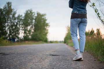 Woman legs in the shoes and jeans on the countryside road. Person waits the bus or a car.