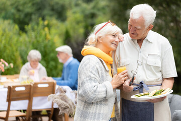Couple of cute seniors standing close to each other and hlding plate full of food during garden...