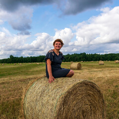 Naklejka na ściany i meble Woman on a mown field in summer, posing next to a roll of hay.