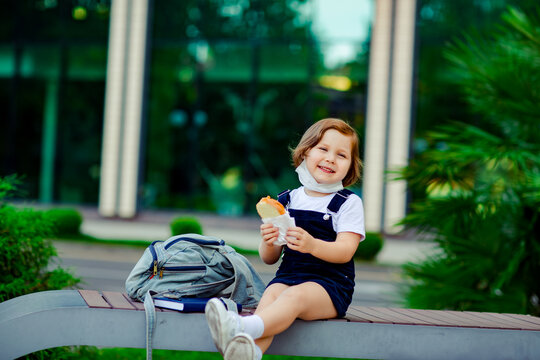 a little schoolgirl, near the school, is sitting on a bench and eating a sandwich, a medical mask on her face
