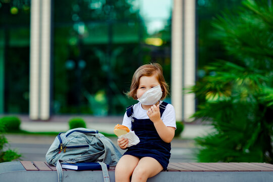 a little schoolgirl, near the school, is sitting on a bench and eating a sandwich, a medical mask on her face