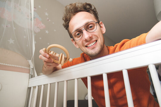 Young Happy Caucasian Father Looking At His Baby In Crib Shaking With Toy.