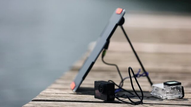 An action camera  is charged from a small solar panel  against the background of a lake at the wooden pier.