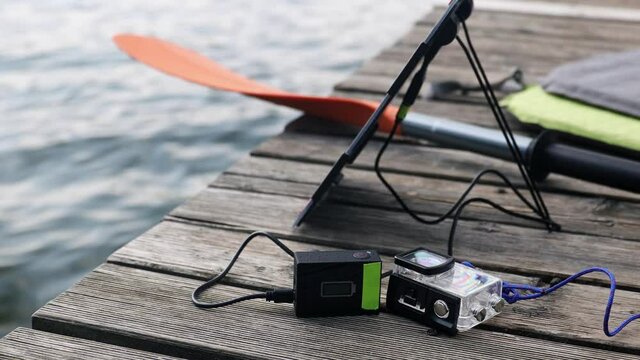 An action camera  is charged from a small solar panel  against the background of a lake at the wooden pier.