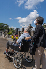 Naklejka premium 車椅子に乗った夫を介助しながらお盆の墓参りに向かうマスクをした老夫婦。暑い日差しの照り付ける夏の日に、花を持って墓地を進む（縦）