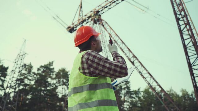 Caucasian engineer in protective uniform and orange hard hat holds walkie-talkie and clipboard on background of electric transmission line. Electrical worker with radio set near high voltage tower.