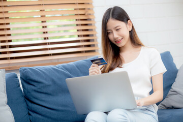 Young asian woman sit on sofa using laptop computer shopping online with credit card buying to internet, happy girl payment with e-business on couch, purchase and payment, business concept.