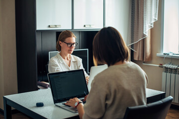 Two young adult women sitting at table facing each other, working on laptops in the home interior. Focus on face of blonde with glasses. Freelance, coworking, remote work, self-employment.