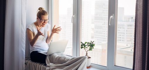 Happy smiling woman wearing glasses sitting on windowsill at city apartment, resting, looking at webcam on laptop, talking on video chat with her friends or family.