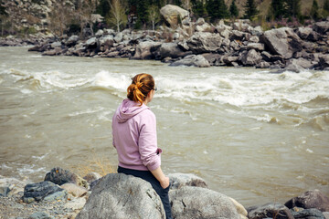 Lonely young brunette woman in sad mood sitting back to the camera on big grey stone near stormy mountain river.