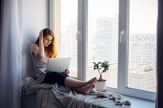 Beautiful positive girl using laptop, sitting on the windowsill in city apartment. Young red-haired woman working at home. Freelance concept.