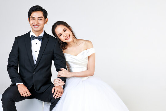 Young attractive Asian couple, soon to be bride and groom, woman wearing white wedding gown. Man wearing black tuxedo, sitting down together. Concept for pre wedding photography