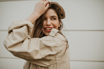 Pretty young woman in beige trench coat smiles sincerely, ruffles hair, looks away and poses...
