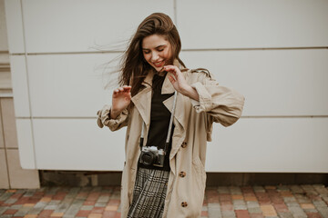 Happy young woman in stylish checkered skirt, black top and trench coat smiles sincerely, looks...