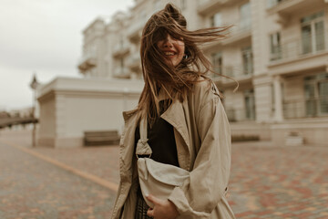 Fototapeta premium Charming woman with fluttering hair smiles outside. Attractive lady in trench coat holds beige handbag and walks.