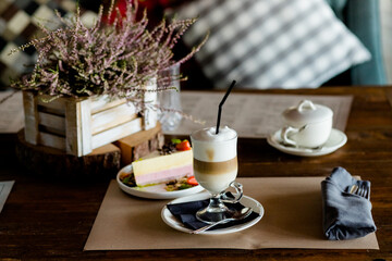 Hot latte macchiato coffee with tasty foam clear glass on dark wooden table serving with sweets, strawberry cake and cane sugar. Breakfast time. Retro toned image. Low key dark photo.