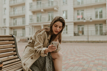 Fototapeta premium Cheerful brunette woman in grey checkered skirt and long beige trench coat chats in cellphone and sits on wooden bench outside.
