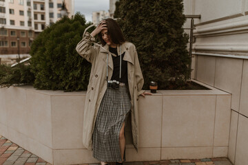 Portrait of long-haired attractive woman in midi plaid dress, black top and beige trench coat leans...