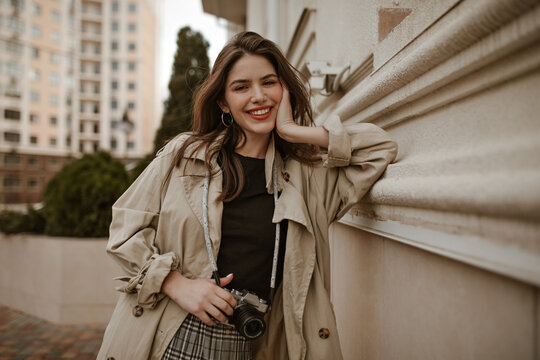 Cool young attractive woman in beige trench coat and black top leans on wall outside, smiles and holds retro camera.